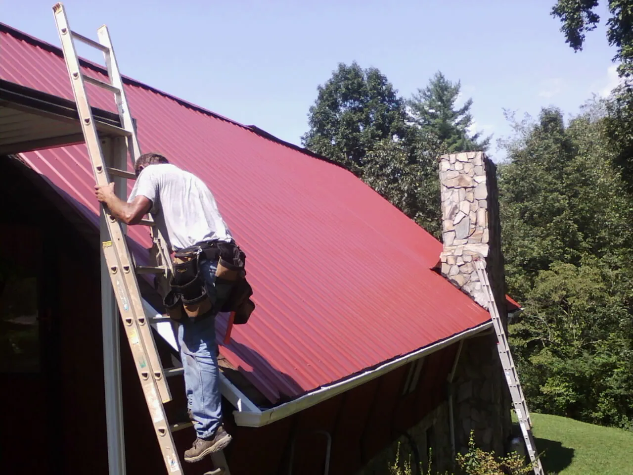 Roofer on a ladder beside a red metal roof and stone chimney — Soffit & Fascia Repair by Roofing Brothers in Merrimack